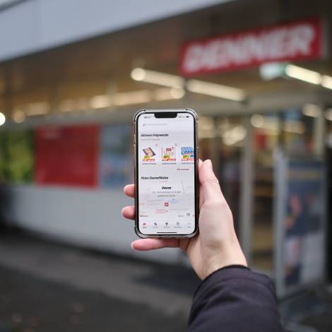 Handy mit geöffneter Denner App in der Hand einer Person vor Denner Filiale